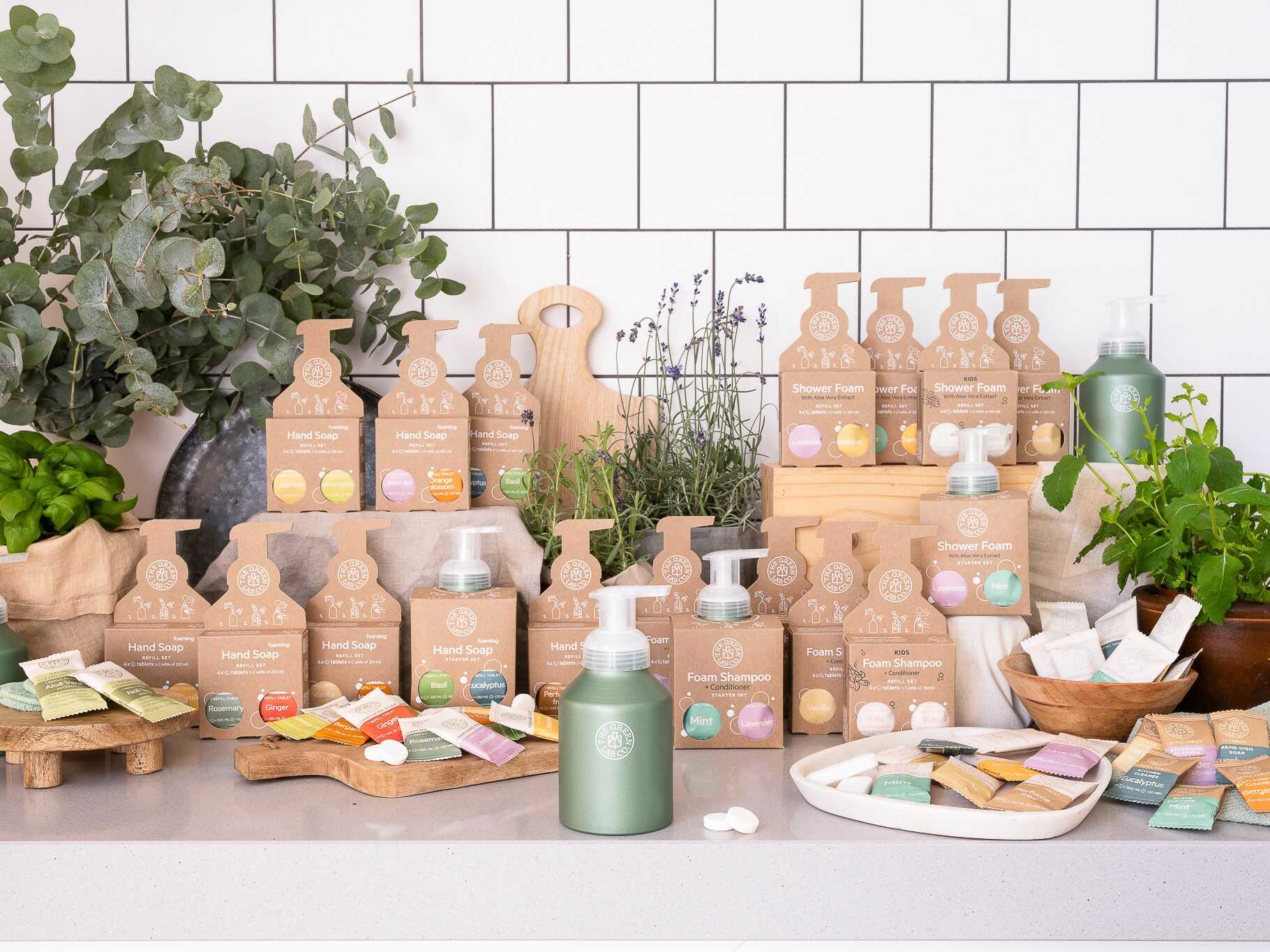 Display of The Green Lab Co refill products—foam hand soap, shower foam, and foam shampoo tablets—with reusable aluminum pump bottles and botanicals on a tiled kitchen backdrop